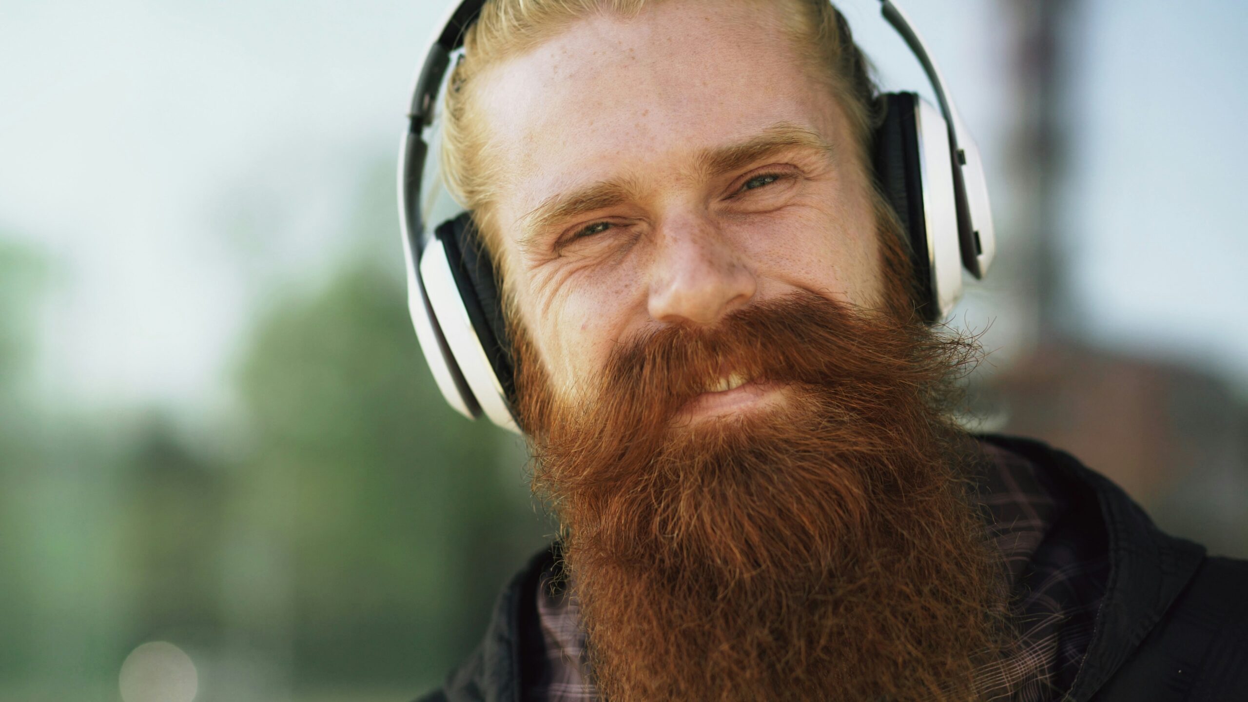Closeup portrait of young bearded hipster man with headphones listen to music and smiling on city street