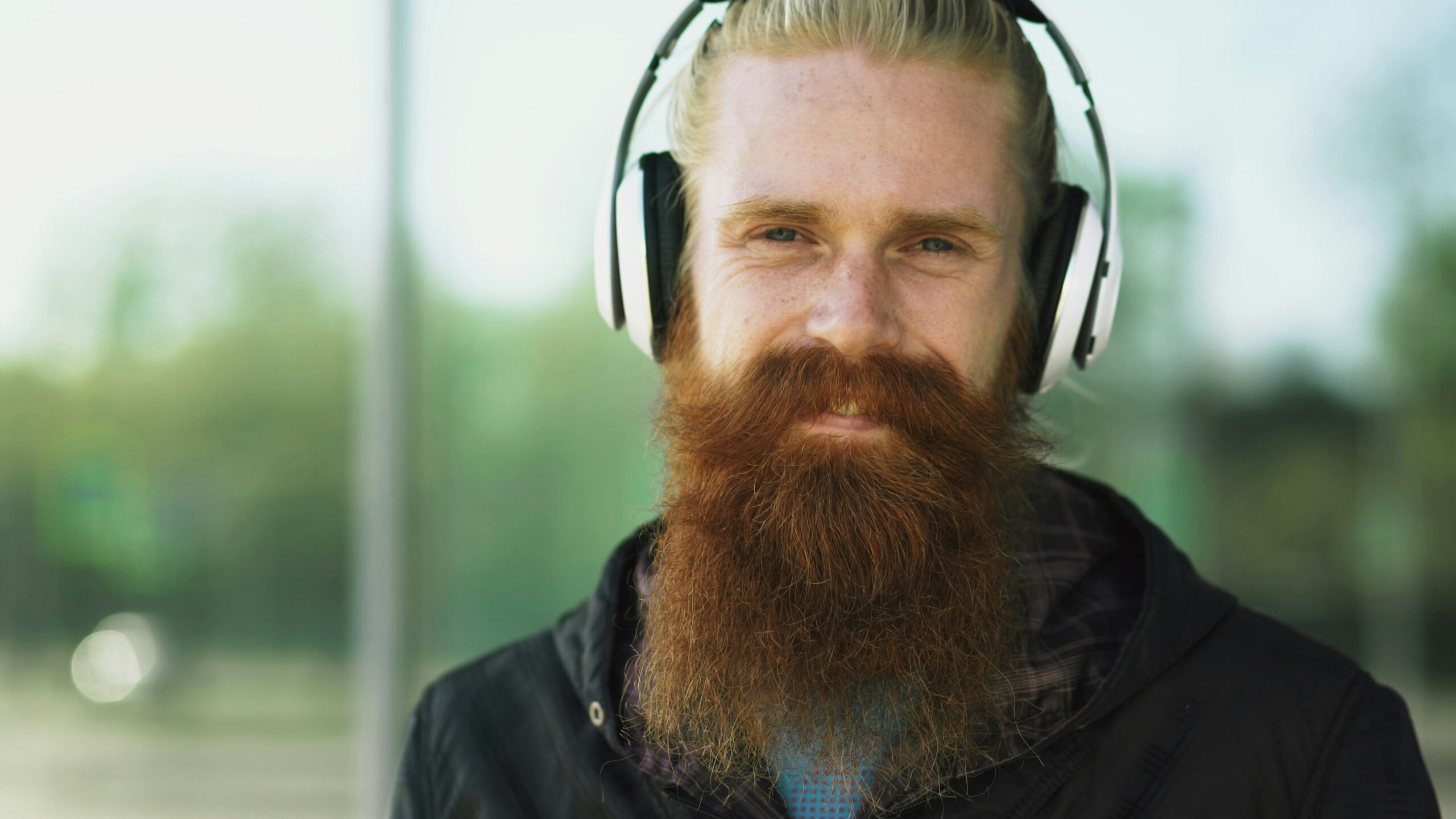 Closeup portrait of young bearded hipster man with headphones listen to music and smiling on city street