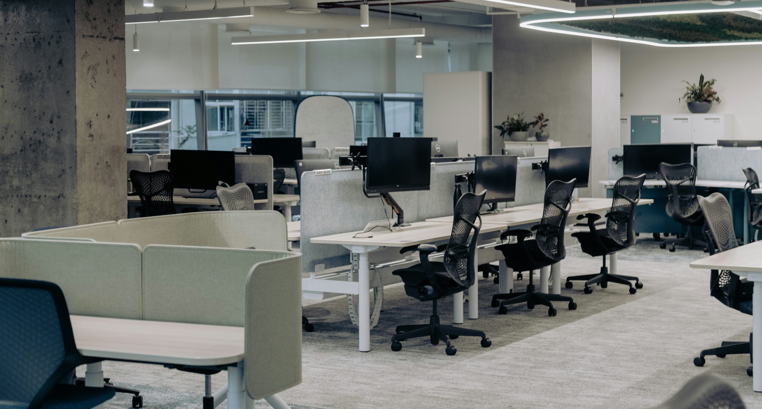 The image depicts a modern, open-plan office with a clean and organized layout. The workspace features multiple desks equipped with computers and ergonomic chairs. The desks are separated by low dividers, creating individual workstations while maintaining an open and collaborative environment. Overhead lighting and large windows provide ample illumination, enhancing the bright and airy atmosphere of the office. The decor includes minimalistic elements with a few potted plants adding a touch of greenery. The overall design emphasizes functionality and comfort, making it a conducive environment